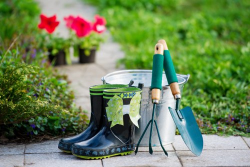This image depicts a garden scene with a paved pathway bordered by lush green grass and various flowering plants. In the foreground, there is a pair of black rubber gardening boots with green and yellow patterned socks hanging over the top, and beside them, a white plastic bucket with gardening tools such as a trowel and transplanter resting inside. The background features vibrant pink flowers in pots, situated along the pathway, with dense greenery creating a natural, well-maintained outdoor space. The scene suggests an active gardening environment, typical of a residential garden in Rotherhithe, with a focus on outdoor plant care, lawn maintenance, and garden preservation activities offered by companies like Gardening Services Rotherhithe.