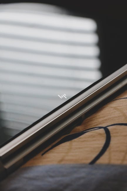 The image shows a close-up of a sleek silver laptop resting on a wooden surface, partially open with the HP logo visible on the lid. The scene is set indoors with blurred horizontally striped window b