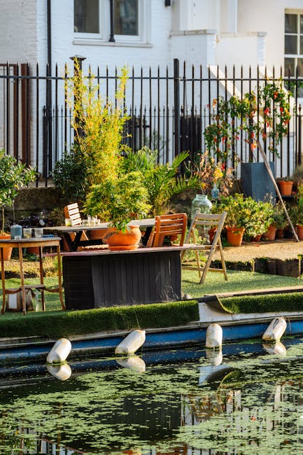 A well-maintained urban garden adjacent to a white residential building, featuring a small pond with floating aquatic plants and a floating barrier with white spheres. The garden has a narrow strip of